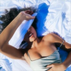 A woman lies on white sheets, face partially covered, in a dreamy setting.